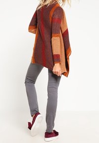 Pletený pončo v oranžové, hnědé a vínové barvě s pruhovaným vzorem; nošené přes šedé kalhoty a vínové boty. Ležérní, volný střih.