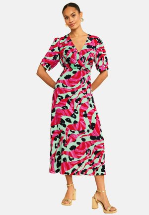 Nő élénk, piros, fekete és mentazöld absztrakt mintákkal díszített színes midi ruhában, arany magas sarkú szandállal párosítva.