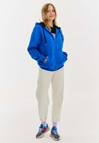 Niebieska bluza z kapturem na zamek błyskawiczny, z czarnym obszyciem kaptura, połączona z jasnobeżowymi spodniami i wielokolorowymi sneakersami. Posiada kieszenie i luźny fason.