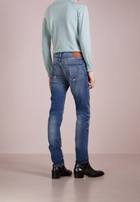 Světle modrý top s dlouhým rukávem, modré slim-fit džíny s odřením, hnědá kožená visačka na pase a černé kotníkové boty s nízkým podpatkem.