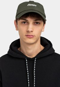Gorra verde oscuro con el texto "Element" en blanco, visera curva y correa ajustable, llevada por una persona con una sudadera negra con cordones estampados.
