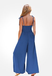 Mono azul con cuerpo fruncido, tirantes anudados, diseño de pierna ancha y bolsillos. Ligero y de textura suave, adecuado para el clima cálido.