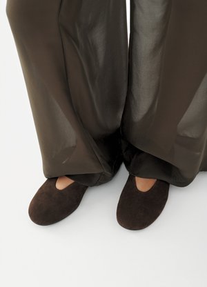 Persoană care poartă papuci din piele întoarsă maro închis cu o mică tăietură, asortați cu pantaloni largi, semi-transparenți, maro închis care acoperă gleznele.