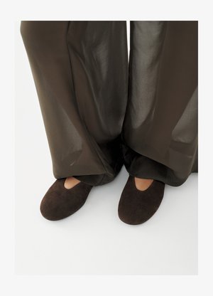 Person, die dunkelbraune Wildlederschlappen mit einer kleinen Aussparung trägt, kombiniert mit weiten, halbtransparenten dunkelbraunen Hosen, die die Knöchel bedecken.