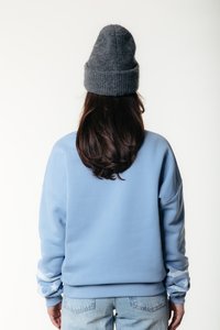 Person med langt brunt hår iført en grå strikket lue, en lyseblå hettegenser med stjernemønster på ermene, og lyseblå jeans, vendt bort.