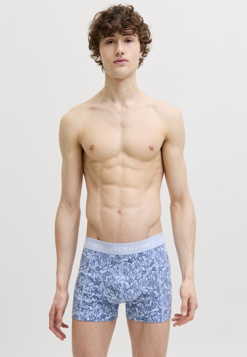 Jonge man met krullend haar staat tegen een effen achtergrond en draagt lichtblauwe boxer shorts met een bloemmotief.