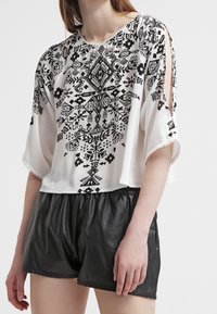 Blouse blanche à manches courtes avec motifs tribaux noirs, design court et détail fendu aux manches, associée à un short en cuir noir taille haute.
