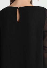 Svart klänning med öppen ryggslut, med en slät textur och genomskinliga ärmar, som betonar elegans och mångsidighet i designen.