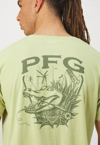 Tricou din bumbac de culoare verde deschis, având un grafic de culoare verde închis cu un aligator înconjurat de plante, cu inscriptionarea "PFG" deasupra designului.