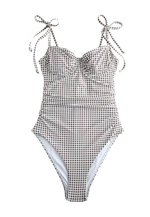 Kvinde i en gingham-farvet badedragt og bredskygget hat, stående barfodet på en sandstrand nær en klippefyldt kystlinje.