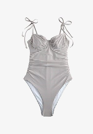 Femeie în costum de baie în carouri, întreg, și pălărie cu boruri largi, stând desculță pe o plajă nisipoasă lângă țărmul stâncos.