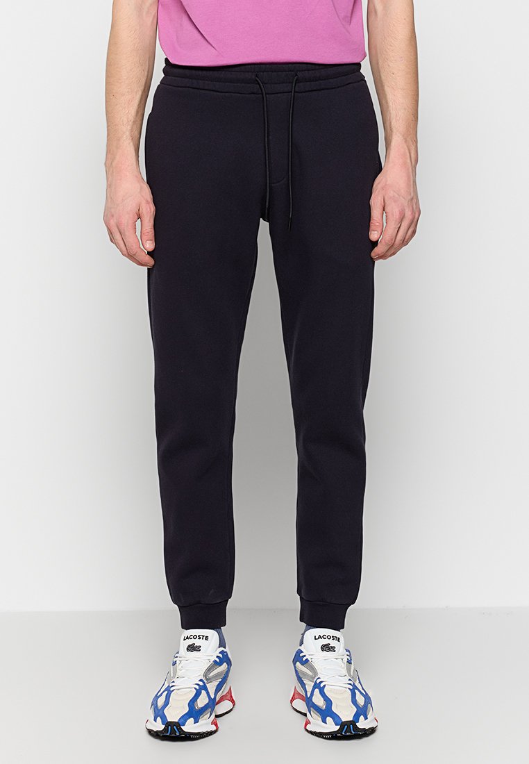 Uomo che indossa pantaloni jogger neri, scarpe da ginnastica Lacoste bianche e blu, e una maglietta rosa, in piedi su un pavimento bianco contro un muro bianco.