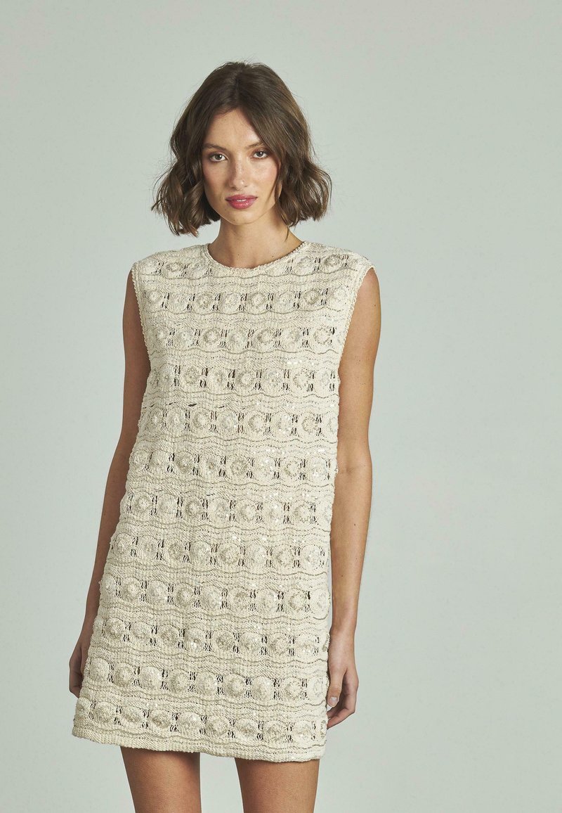 Ärmelloses Strickkleid in Creme mit einem strukturierten Muster, das Reihen von kreisförmigen und wellenförmigen Designs zeigt. Es hat eine kurze Länge und einen runden Halsausschnitt.