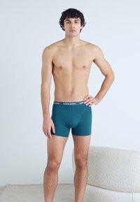 Tirkizne muške bokserice od mekog materijala, s brendiranim elastičnim pojasom i prilagođenim krojem koji ističe tjelesnu konstituciju.