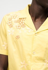 Κίτρινο κοντομάνικο πουκάμισο με κουμπιά και κεντητά πορτοκαλί και λευκά λουλουδένια σχέδια στο στήθος και στα μανίκια, που φοριέται από ένα άτομο με σκούρο δέρμα.