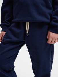 Person, der bærer marineblå joggingbukser med hvid snor og matchende marineblå sweatshirt, hænder afslappet ned langs siderne.