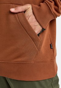 Pullover marrone con tasca a marsupio e polsini e orlo a costine. Il tessuto è morbido con una superficie leggermente strutturata. Accentuato con un cordino.