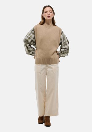 Un gilet sans manches beige sur une chemise à carreaux verte, associé à un pantalon large crème et des bottines brunes, se tenant devant un fond blanc.