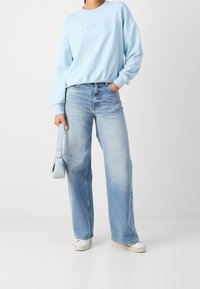 Light blue sweatshirt with embossed text, paired with high-waisted, wide-leg faded blue jeans and white footwear, holding a light blue bag.