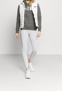 Gilet bianco e gonfio sopra un felpa grigia con cappuccio, abbinato a leggings aderenti di un grigio chiaro e scarpe da ginnastica bianche. Texture morbide e colori neutri predominano.