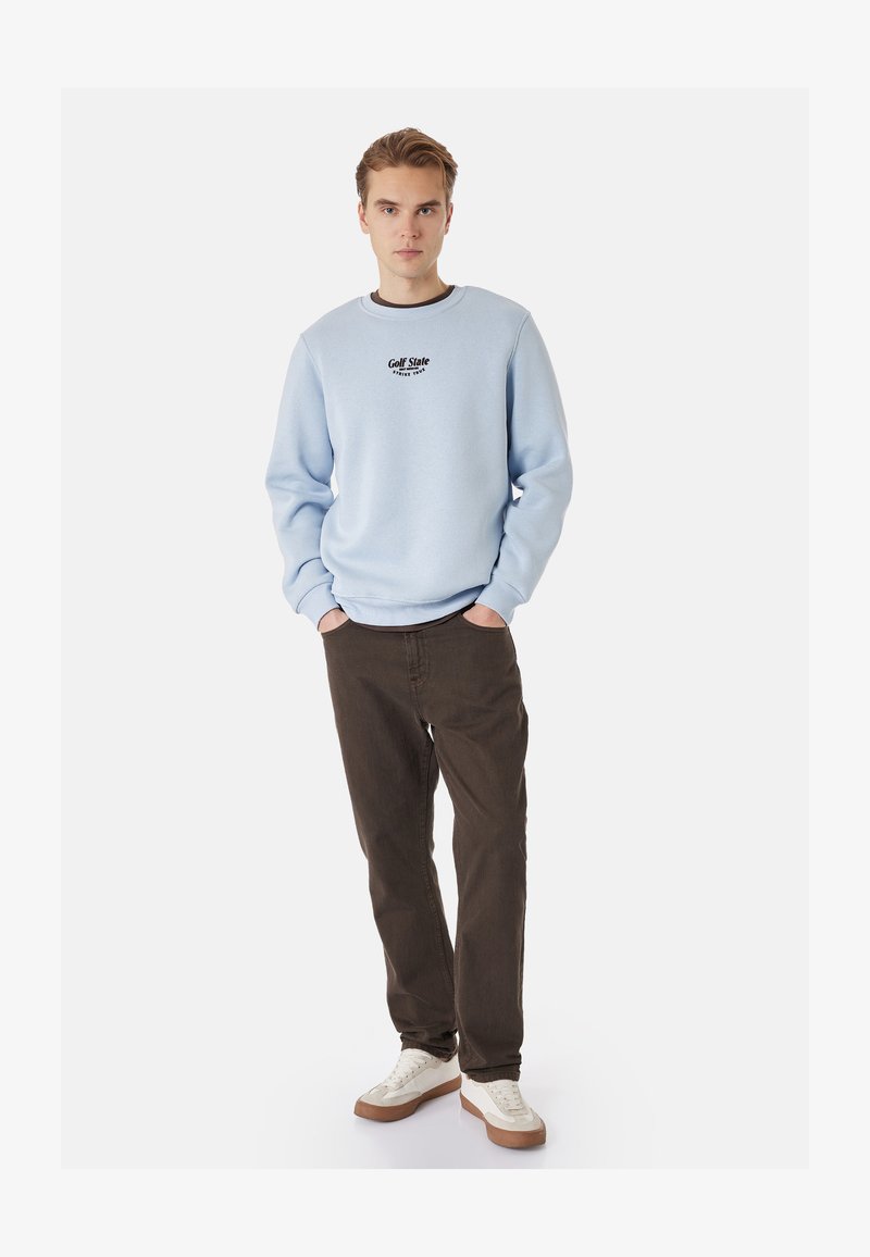 Jeune homme debout, les mains dans les poches, portant un sweat-shirt bleu clair, un pantalon marron foncé et des baskets blanches sur un fond uni.