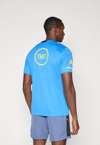 Zila sporta krekls ar īsām piedurknēm, kas aizmugurē rotāts ar dzeltenu apļa logo ar uzrakstu "THFC", un dzeltenu tekstu "Alipay" uz piedurknes.