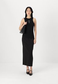 Čierny top bez rukávov s logom, skombinovaný s dlhou čiernou sukňou s pásikmi a čiernymi topánkami s špicatou špičkou. Model drží čiernu kabelku.