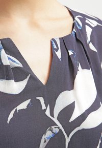 Tessuto blu navy con grandi motivi a foglia bianchi e blu. Scollatura a V con pieghe al centro, che creano un effetto drappeggio sottile.