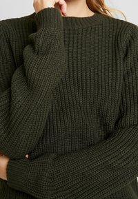 Maglione verde lavorato a maglia con texture a coste, scollatura rotonda e maniche lunghe. Il materiale sembra morbido e spesso, adatto per il clima più freddo.