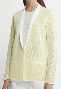 Blazer en maille jaune clair avec revers blancs et fermeture à boutons. Comprend deux poches avant et un tissu texturé et respirant.