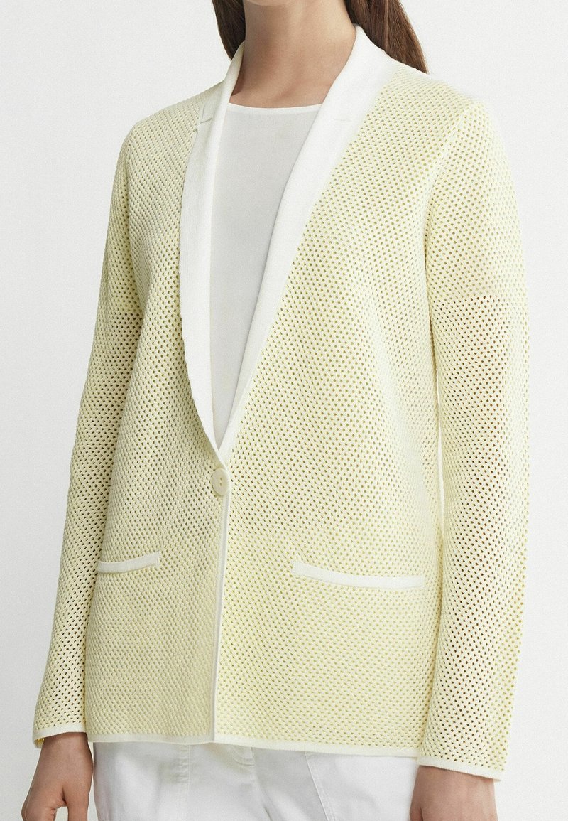 Blazer en maille jaune clair avec revers blancs et fermeture à boutons. Comprend deux poches avant et un tissu texturé et respirant.