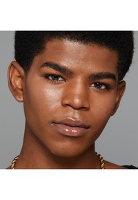 Primo piano di un volto con pelle liscia di un marrone chiaro, capelli ricci scuri e occhi chiari. Presenta labbra lucide e una collana di catena d'oro.