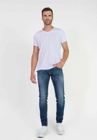Camiseta de algodón blanca con cuello en V, jeans de mezclilla ajustados azules y zapatillas blancas, de pie contra un fondo liso. Diseño de atuendo casual.