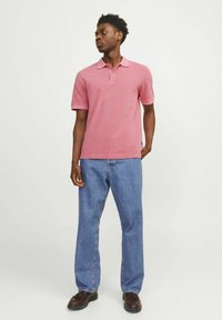 Polo majica s kratkimi rokavi, svetlo roza barve, z ovratnikom, v kombinaciji z ohlapnimi modrimi jeanskimi hlačami in temno rjavimi čevlji.