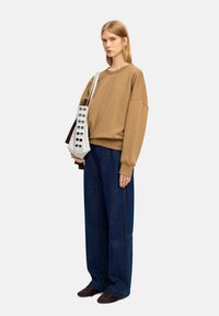 Brązowy oversize sweatshirt z opuszczonymi ramionami, niebieskie szerokie dżinsy i ciemnobrązowe buty. Modelka trzyma szary worek z okrągłymi wycięciami.