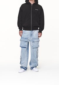 Felpa con zip nera con tasche frontali. Pantaloni cargo azzurri chiaro con diverse tasche laterali e vestibilità ampia. Scarpe da ginnastica bianche.