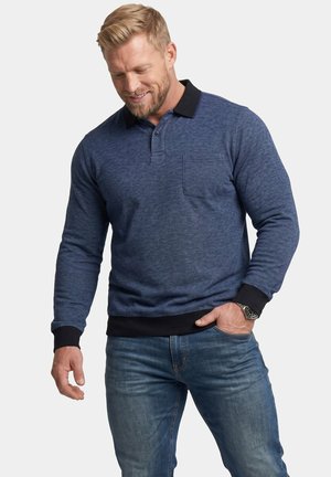 Mand iført en langærmet blå polo med sort krave og manchetter og blå jeans, smilende og kiggende nedad.