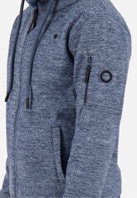 Niebieska heatherowa bluza z kapturem na zamek, z troczkami, boczną kieszenią, kieszenią na zamek na rękawie oraz okrągłą czarną naszywką na górnej części ramienia.