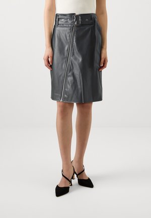 Jupe en cuir noire mi-longue avec fermeture éclair diagonale à l’avant et large ceinture, portée avec des talons noirs pointus et un haut blanc sans manches.