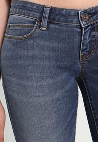 Pantalones vaqueros de mezclilla azul oscuro con un diseño ajustado, que presentan un bolsillo delantero, costuras cosidas y un cierre de botón metálico. Textura suave.