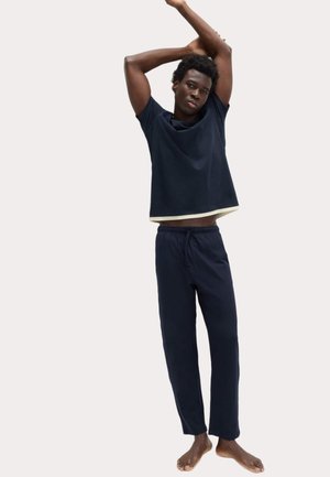 Jeune homme en tenue de détente bleu marine, s'étirant avec les bras levés au-dessus de la tête, devant un fond clair uni.