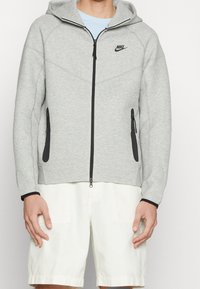 Harmaa vetoketjutakki hupulla, mustilla yksityiskohdilla ja pehmeällä pinnalla. Siinä on sivutaskut ja Nike-logo rinnassa.