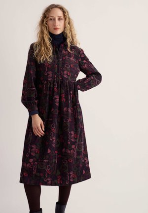 Woman with curly blonde hair wearing a long-sleeve, dark patterned dress with burgundy tights and black boots against a plain background.