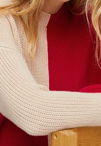 Sweter z dzianiny z podzielonym wzorem, z teksturowanym beżowym lewym rękawem i gładką czerwoną częścią główną, prezentujący kontrastujące kolory i materiały.