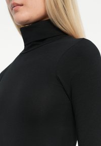 Černý rolák s přiléhavým střihem, vyrobený z hladké látky. Má vysoký límec a dlouhé rukávy, s čistými, minimalistickými liniemi.