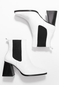 Botas de tobillo de cuero blanco con paneles laterales elásticos negros, puntera afilada y tacón grueso negro. Cuenta con una lengüeta para facilitar el uso.
