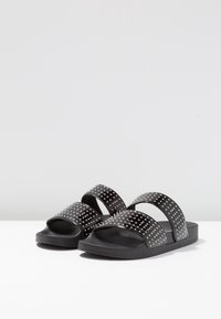 Des sandales noires à glissière présentent deux larges sangles ornées de clous en argent, en matériau texturé, et une semelle plate pour le confort.