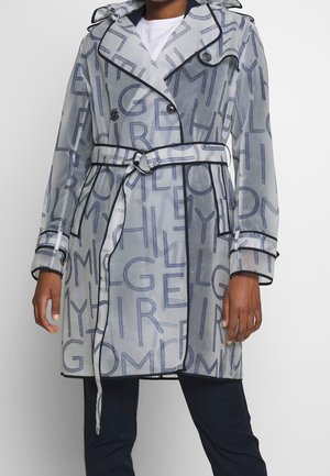 Woman wearing a sheer, belted trench coat with large printed letters and dark trim over a white top and dark pants.