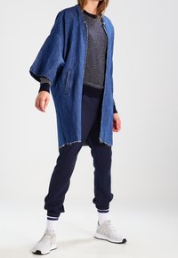 Denim long coat with raw edges, worn over a navy polka-dot sweater and navy joggers. Finished with white-accented athletic shoes.
