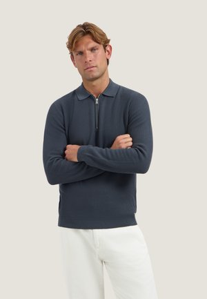 Hombre con cabello castaño claro, vestido con un suéter gris oscuro con cremallera y pantalones blancos, está de pie con los brazos cruzados frente a un fondo claro y sencillo.